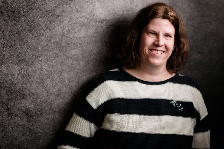 Smiling woman with brown hair in front of a grey wall, natural portrait, friendly appearance.