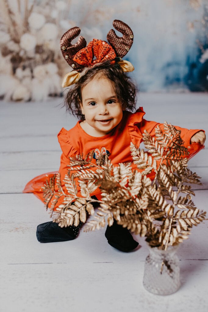 Fröhliches Kind in festlichem Weihnachten-Outfit mit Rentier-Haarreif, auf Holzboden sitzend, schön dekorierter Hintergrund, professionelle Fotografie, Kinderfotostudio München, festliche Kinderfotos, Weihnachtsbilder, kindgerechte Porträts, Shooting für Familien und Kinder in München.