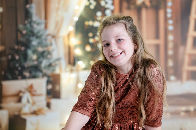 Weißer Hintergrund, festliches Dekor, junge Frau in rotem Pailletten-Kleid lächelt, Weihnachtsbaum im Hintergrund, warme Beleuchtung, festliche Stimmung, Portraitaufnahme, festliche Kleidung.