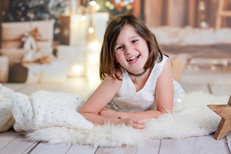 Porträt eines fröhlichen jungen Mädchens, das auf einem weichen weißen Fell liegt, mit Weihnachtsdekoration im Hintergrund, festliche Stimmung, kreative Familienfotografie in München.