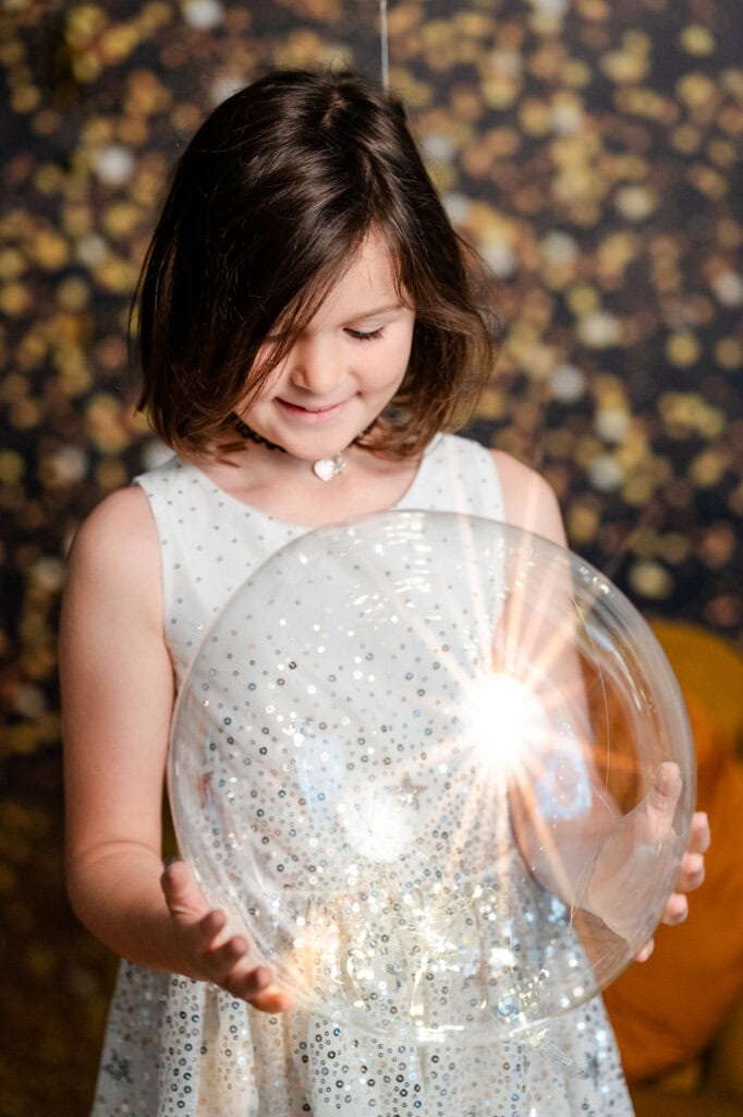 Blondes Mädchen in weißem Kleid mit Glitzer und einem Herzschmuck, das einen durchsichtigen Ballon mit Lichter glänzend hält. Der Hintergrund ist dunkel mit warmen, goldenen Flecken, die eine festliche Atmosphäre schaffen.