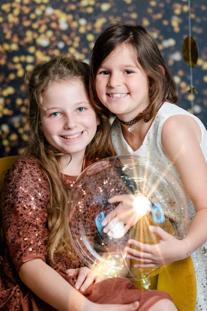 Zwei fröhliche Mädchen in festlichen Outfits halten eine leuchtende Weihnachtskugel vor gold-funkelndem Hintergrund im Fotostudio Photogenika.