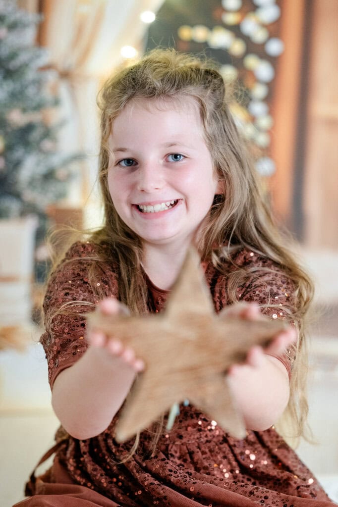 Fröhliches Mädchen mit goldbraunen, lockigen Haaren, das eine holzfarbene Sternfigur zum Spielen hält, vor einem weihnachtlich dekorierten Hintergrund.