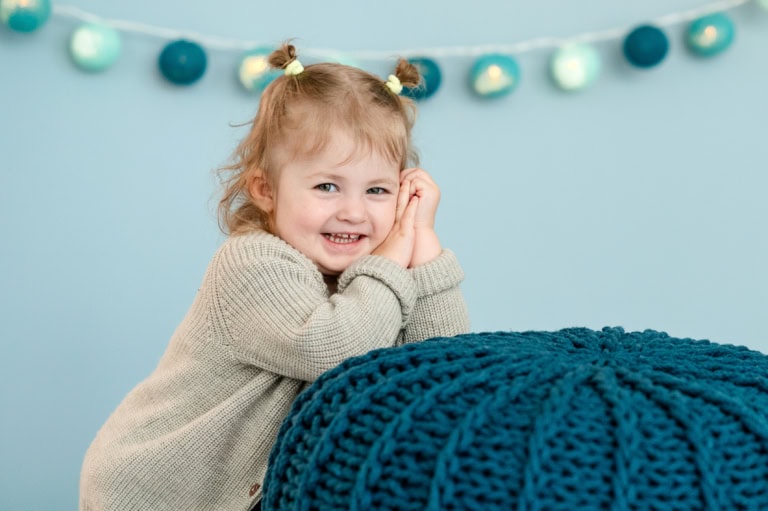 Ein glückliches Kleinkind lehnt an einem blauen Strickpouf.