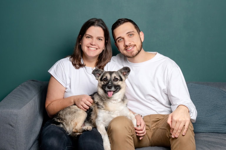 Lächelndes Paar mit Hund auf Sofa vor grüner Wand, professionelle Familienfotografie, emotionale Momentaufnahme, Portraitfoto, München, Fotostudio Photogenika.