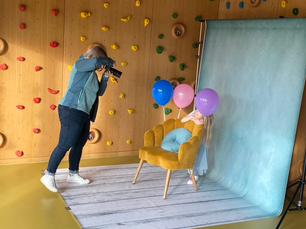 Ein kleines Mädchen mit Ballons sitzt auf einem gelben Sessel vor einem Fotohintergrund, während eine Fotografin sie für ein professionelles Fotoshooting in einem Kindergarten in München fotografiert.