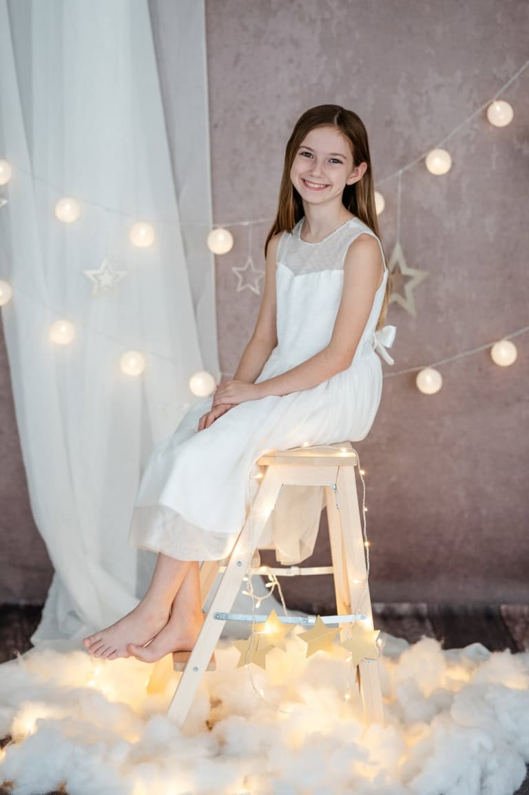 A smiling girl in a white dress sits on a wooden chair in front of a festive decoration with fairy lights and stars.