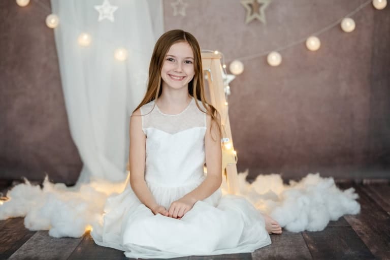 Smiling girl in a white dress sits on a white carpet in front of Christmas decorations.