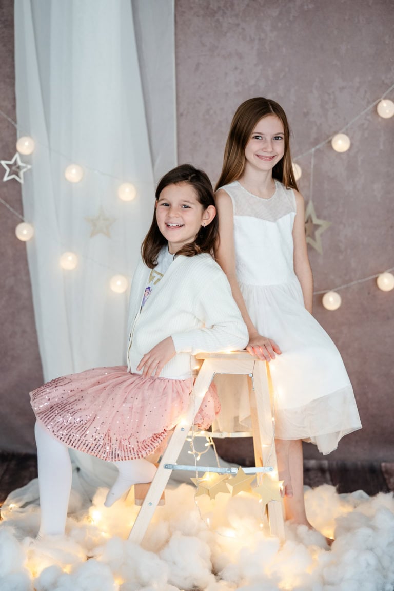 Two laughing girls in white dresses during a festive photoshoot with fairy lights and star decorations.