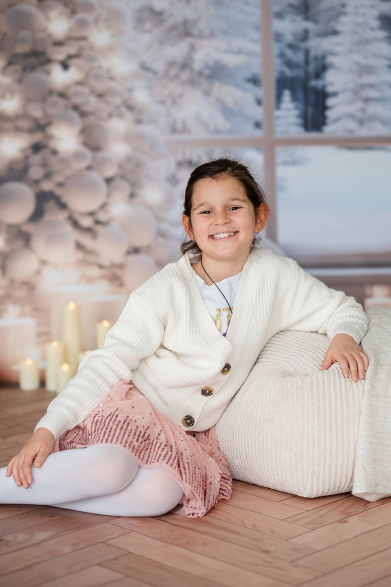 Cheerful girl sitting on a carpet in front of a Christmas tree with candles, winter landscape in the window in the background.