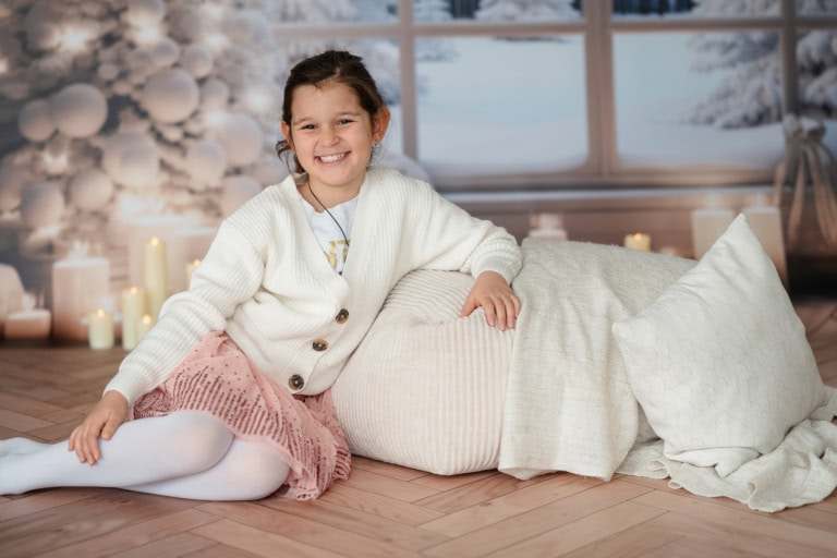 A cheerful girl sits on the floor in front of a wintry window with candles and decorations.