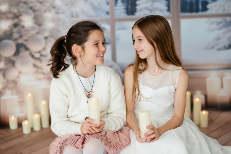 Two laughing girls sit in a festively decorated room with candles and snowflakes in the background.