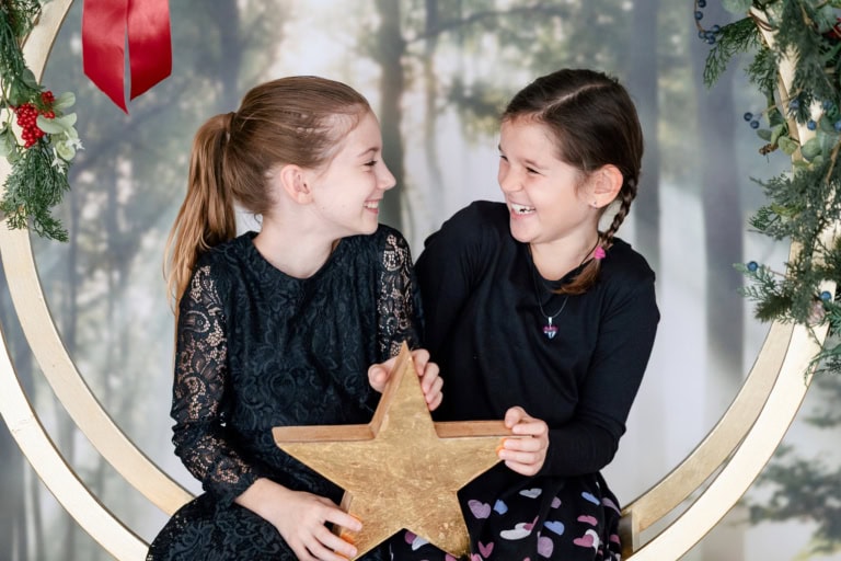 Two laughing girls sit on a Christmas ring, holding a golden star and looking happily at each other.