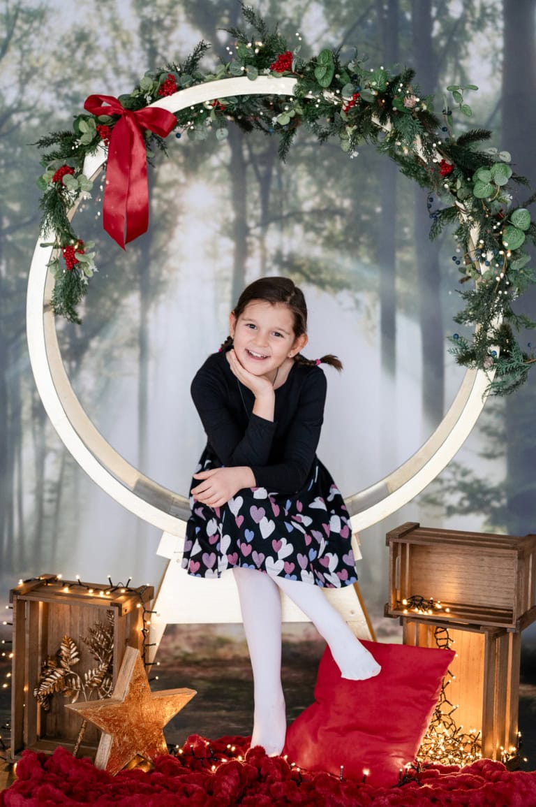 A young girl sits in front of a festively decorated frame with Christmas decorations, smiling and posing for a Christmas photo.