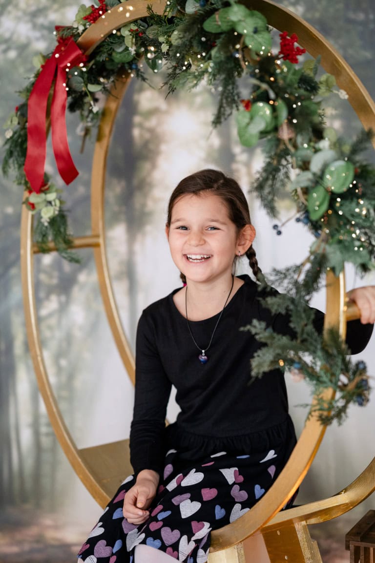 A cheerful girl sits in a golden frame with Christmas decorations, smiling and wearing a black top and trousers with heart patterns.