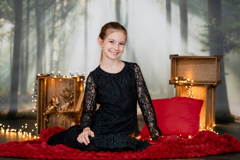 Young woman in black lace dress sits on red carpet in front of a Christmas backdrop with fairy lights and decorations.